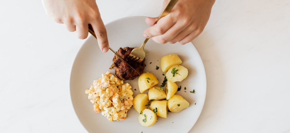 Top view of crop unrecognizable male with cutleries slicing delicious beef cutlet garnished with boiled potatoes and parsley and portion of Russian meat salad on plate placed on white table